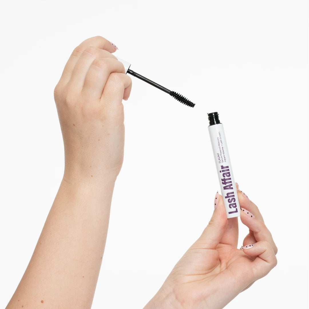 Hand holding a mascara wand above an open tube of 'Lash Affair' mascara on a white background